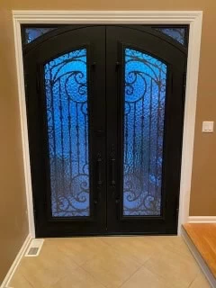 A dark, matte black set of double doors with intricate, ornate wrought ironwork. The doors feature arched tops and are flanked by narrow sidelights, also with decorative ironwork. The glass in the doors and sidelights is textured and opaque, with a prominent blue glow, likely from the lighting on the other side of the doors.