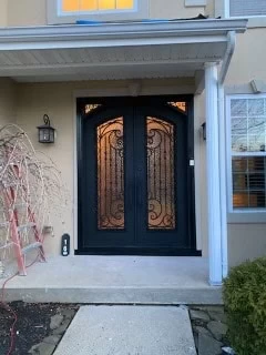 A wide-angle, head-on view of the front entrance of a house. The entrance is a recessed, double-door unit painted matte black. The doors have arched tops and contain two tall, rectangular windows with textured glass and ornate wrought iron scrollwork. The front porch is concrete, and the house has a beige stucco exterior with white trim.