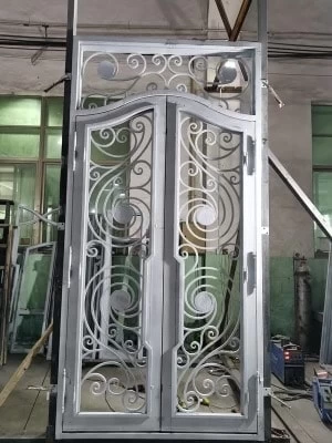 A wide, front-on view of an ornate double-door unit, likely in a workshop. The doors are a light gray primer color, with a delicate and elaborate iron scrollwork design. The design features a mix of tight and loose scrolls, creating a graceful, flowing pattern across both doors.