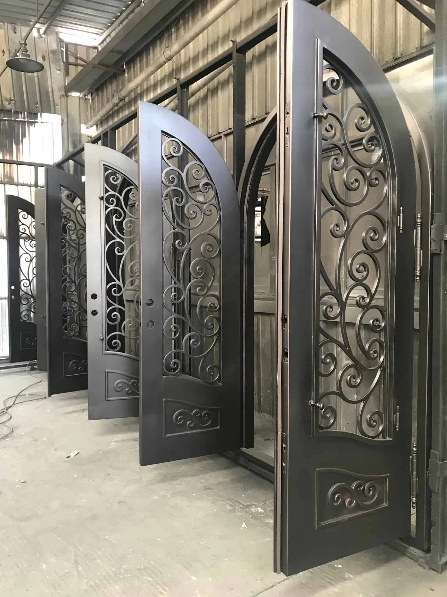 A line of several identical, arched, single wrought iron doors standing in a factory or workshop. The doors are in a dark, unfinished metal state and feature a large, arched window with ornate, swirling scrollwork that fills the space. The scrollwork continues into a rectangular panel at the bottom of the door. The doors are propped open slightly, showing their thickness and a simple, curved handle on the inside.
