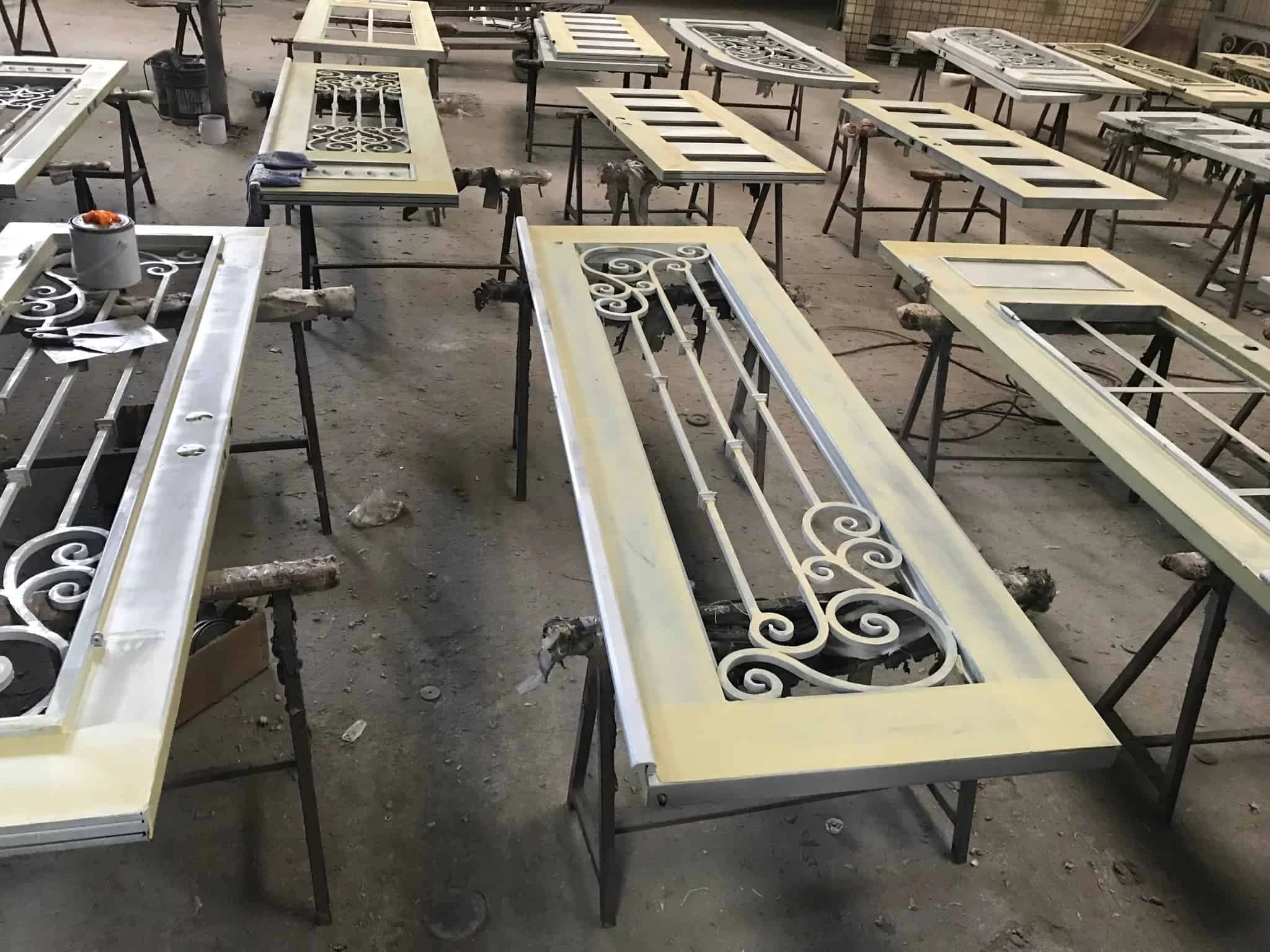 A workshop scene showing several wrought iron doors in various stages of a painting or finishing process. The doors are propped up horizontally on sawhorses. Some doors are a dark gray, while others are coated in a pale, primer-like color. The doors have decorative ironwork and window cutouts. In the foreground, a door with elaborate swirling patterns is being worked on.