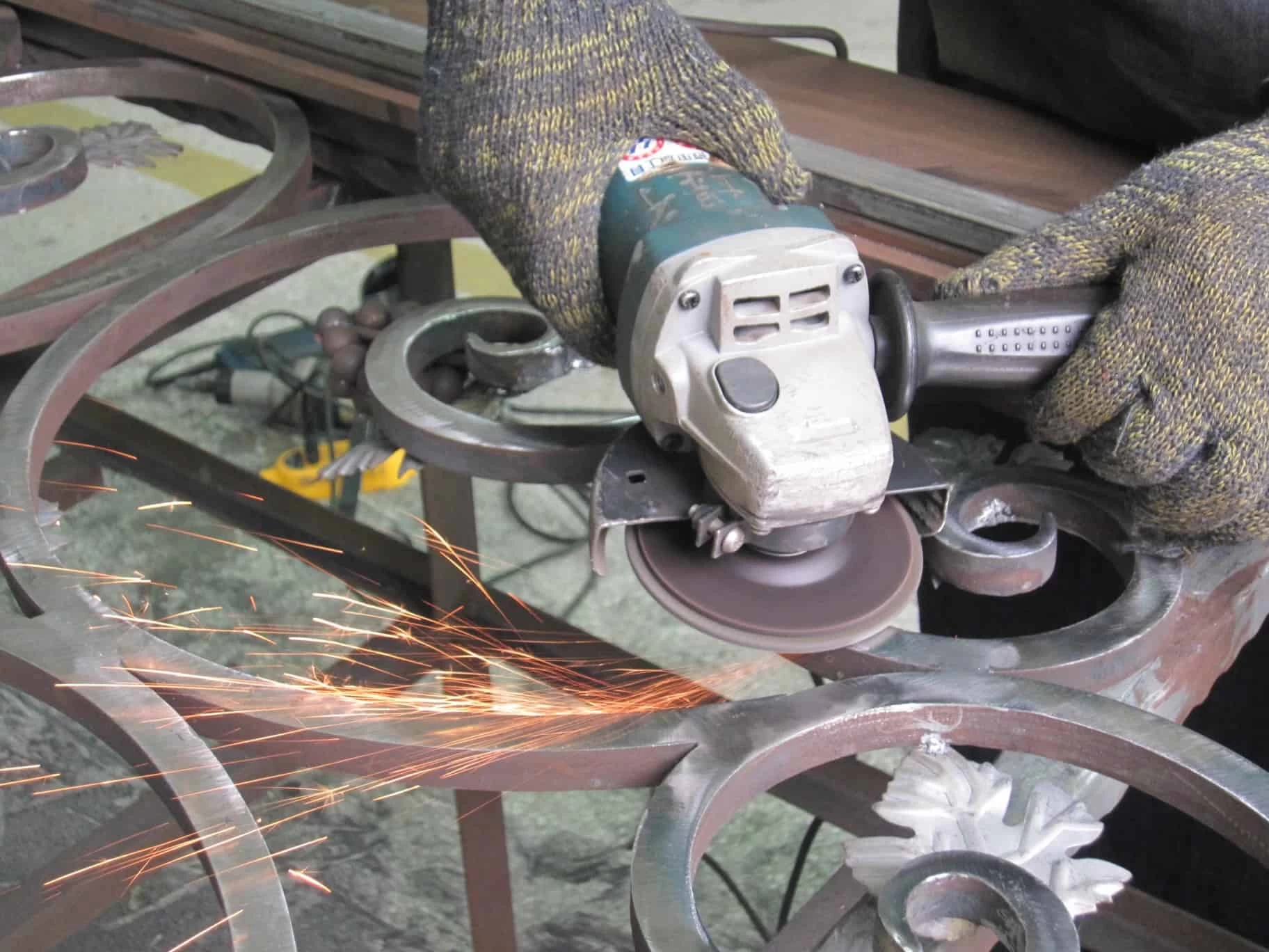 A close-up shot of a worker's gloved hands using an angle grinder on a piece of ornate wrought iron. Sparks are flying from the tool as it grinds against the metal, and the background is out of focus.