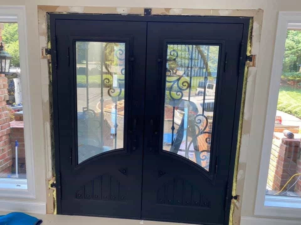 A dark-colored double door is being installed in a new entryway, with the door frame exposed and foam insulation visible. Each door has a tall, arched glass panel with a simple wrought-iron scrollwork design. The bottom of each door has a small decorative panel with a raised triangular detail and vertical lines. The doors are flanked by two large, multi-paned windows, and a brick wall and outdoor lantern can be seen through the window on the left.