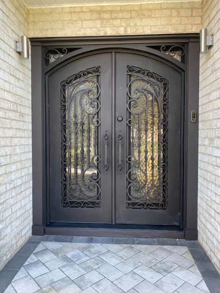 A luxurious, arched double door with a dark bronze finish is set against a light-colored brick facade. The doors feature textured glass panels and intricate, hand-forged wrought-iron scrollwork. Long, matching vertical handles and a simple round lock are positioned at the center. The top corners of the dark door frame are also adorned with complementary scrollwork. The entryway is lit by two modern sconces, and the ground is covered with a herringbone pattern of light gray pavers bordered by darker tiles.
