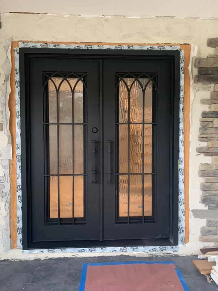 A newly installed set of dark-colored double doors. Each door has a long glass panel featuring a textured, rippled pattern and is covered by a simple, medieval-style arched iron grid. The doors are flanked by a stone veneer wall on the right and a light-colored siding wall on the left. The door frame is exposed, showing a temporary covering, insulation, and the raw wood of the jamb.