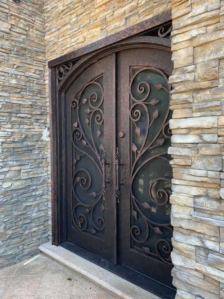 A striking set of grand, arched double doors is set into a wall of stacked stone. The doors have a rich, textured bronze or copper finish and feature intricate, hand-forged wrought-iron scrollwork with subtle leaf details over decorative glass panels. The door handles are long, vertical, and match the ornate design. The door frame is also adorned with complementary scrollwork at the top corners, creating a cohesive, elegant look.