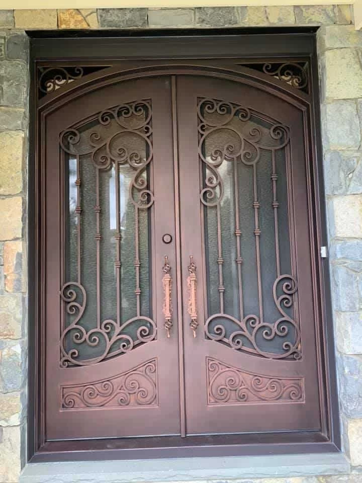 A beautiful set of ornate, arched double doors with a dark, textured bronze or copper finish. Each door has a glass panel behind an intricate, hand-forged wrought-iron scrollwork design. The doors feature two long, vertical handles with a matching finish and a simple round lock at the center. The top corners of the dark door frame are also adorned with complementary scrollwork. The entire entryway is framed by a rustic stacked stone wall.