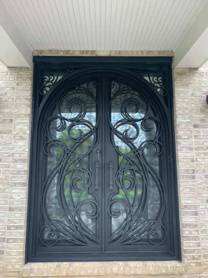 A magnificent set of ornate, arched double doors in a dark, matte finish is set into a light-colored brick facade. The doors feature an intricate, asymmetrical, and flowing hand-forged scrollwork design that covers the large glass panels. The door handles are long and vertical, with a matching ornamental design, and the top corners of the door frame are also adorned with decorative scrollwork.