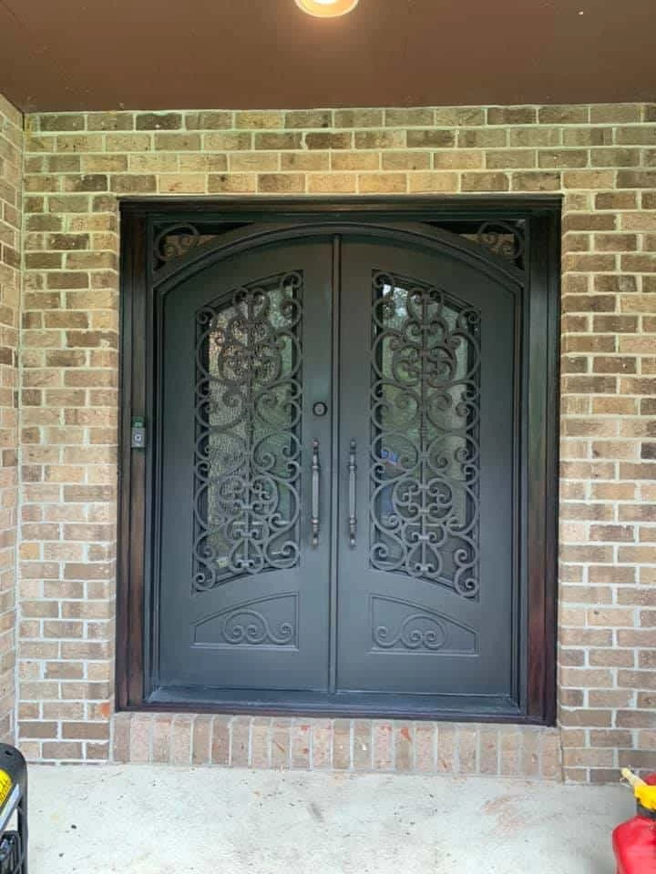 A luxurious set of ornate, arched double doors in a dark, matte finish is set into a red brick facade. The doors feature intricate, hand-forged wrought-iron scrollwork over glass panels. Two long, vertical handles with matching scrollwork are positioned at the center, and a simple round lock is visible above them. A decorative, dark frame surrounds the doors, and the top corners of the frame are also adorned with complementary scrollwork.