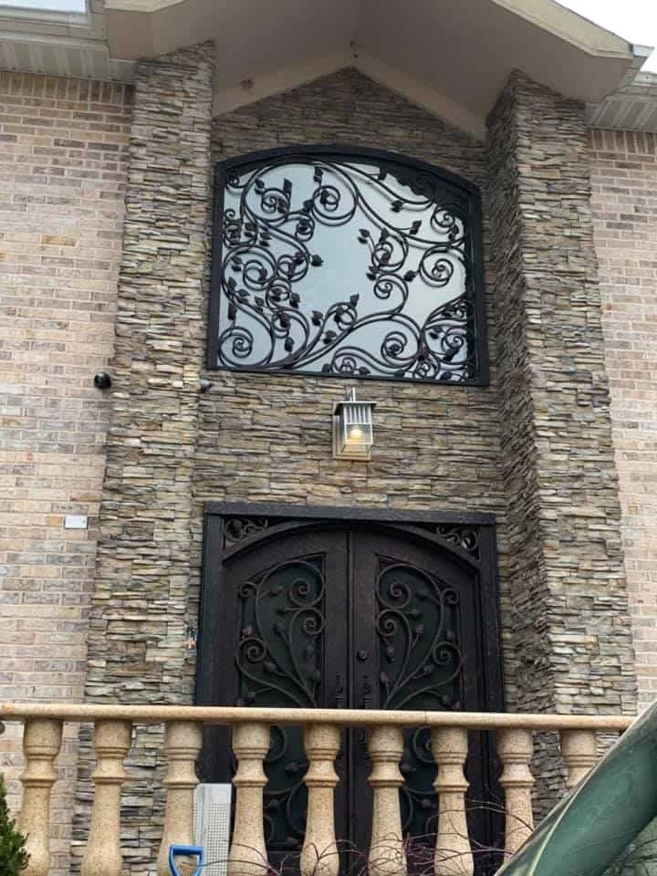 A striking entryway featuring a pair of dark, arched double doors with an intricate wrought-iron scrollwork design over glass panels. Above the doors, a large arched transom window has matching, extensive scrollwork, creating a unified and grand look. The house facade is a mix of light-colored brick and stacked stone veneer. A lantern is mounted between the door and the window, and a stone balustrade is visible in the foreground.