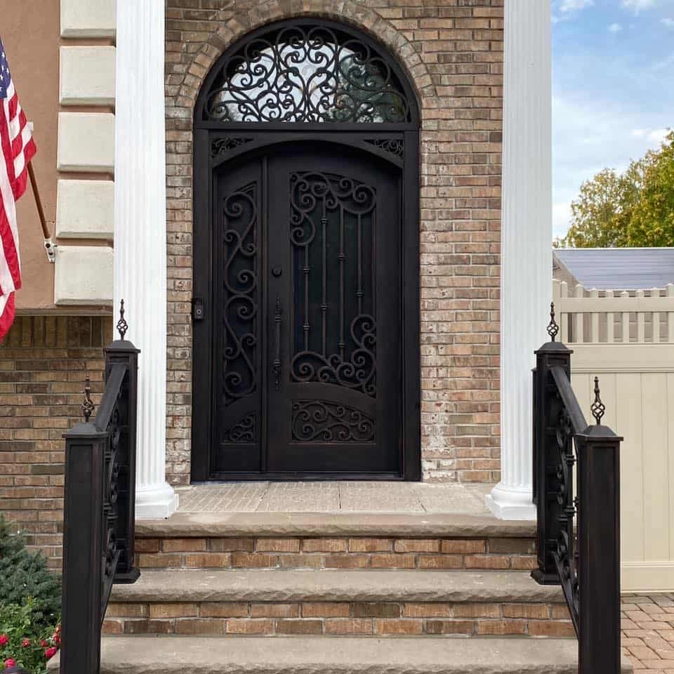 A striking, dark-colored iron front door with an intricate, arched transom window is centered on a brick facade. The door features one full-size panel with ornate wrought-iron scrollwork and a smaller, inactive panel with complementary detailing. The entry is framed by two large white columns and features a brick and stone staircase with dark, decorative iron railings. An American flag is visible on the left side of the house.