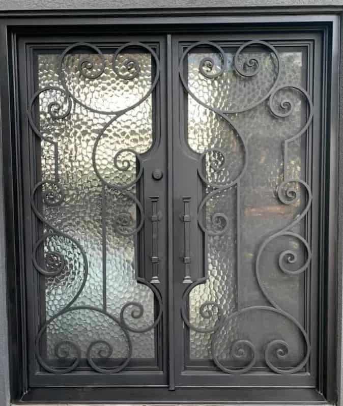 A close-up of a pair of double doors with wrought iron scrollwork and textured, privacy glass panels. The ironwork has large, intricate scrolls that sweep across each door, and the entire frame is painted a matte gray or black.