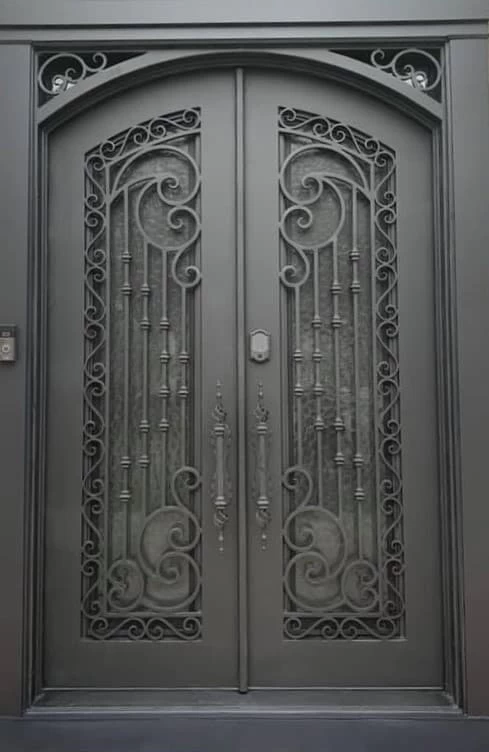A full-frame view of a pair of large, gray double doors with a rounded top. The doors feature a central horizontal lock and vertical handle on each side. The doors have narrow, rectangular window panes with textured glass, each covered by elaborate wrought ironwork with a mix of geometric and swirling patterns. The wrought iron details extend to the arched frame above the doors.