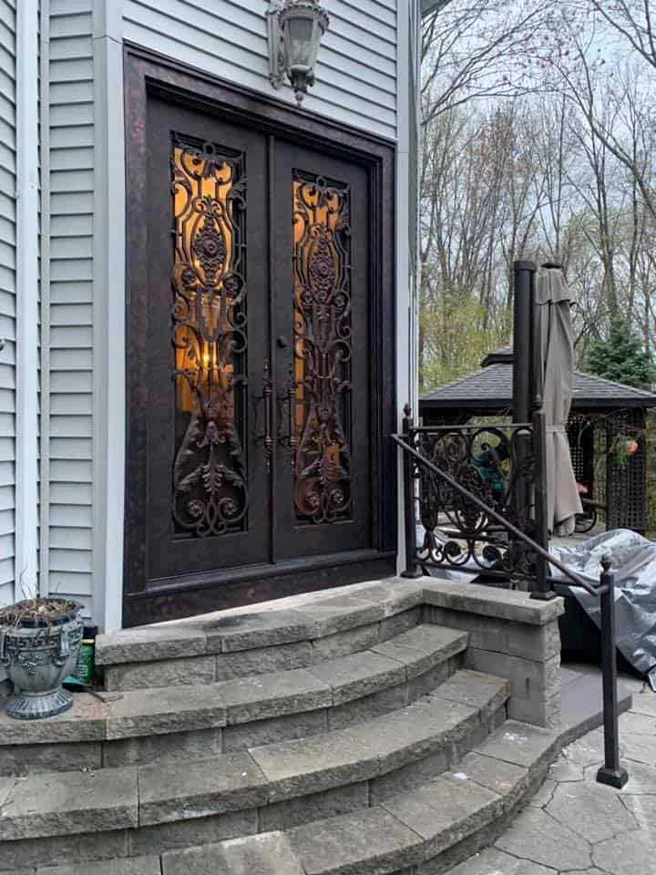 A stunning set of ornate, arched double doors in a dark, textured bronze or copper finish. The doors have decorative glass panels with intricate scrollwork. The entryway features a set of curved stone steps and a black wrought-iron handrail with decorative flourishes. A classic-style lantern hangs on the light gray siding of the house.