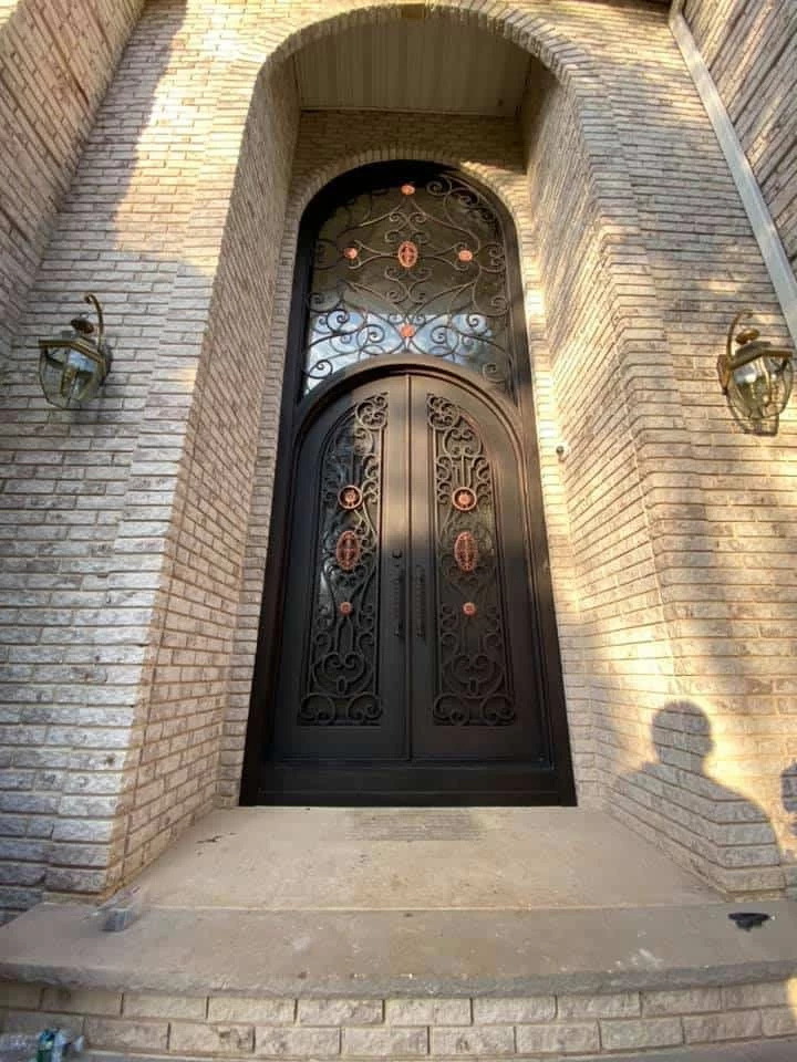 A majestic, dark-colored arched double door is set within a grand, recessed brick entryway. The doors feature intricate, hand-forged scrollwork over glass, accented with oval and circular motifs in a subtle bronze or copper tone. A large, arched transom window above the doors has a matching grid and decorative scrollwork. The brick facade is illuminated by two large, vintage-style lantern sconces mounted on either side of the entrance.