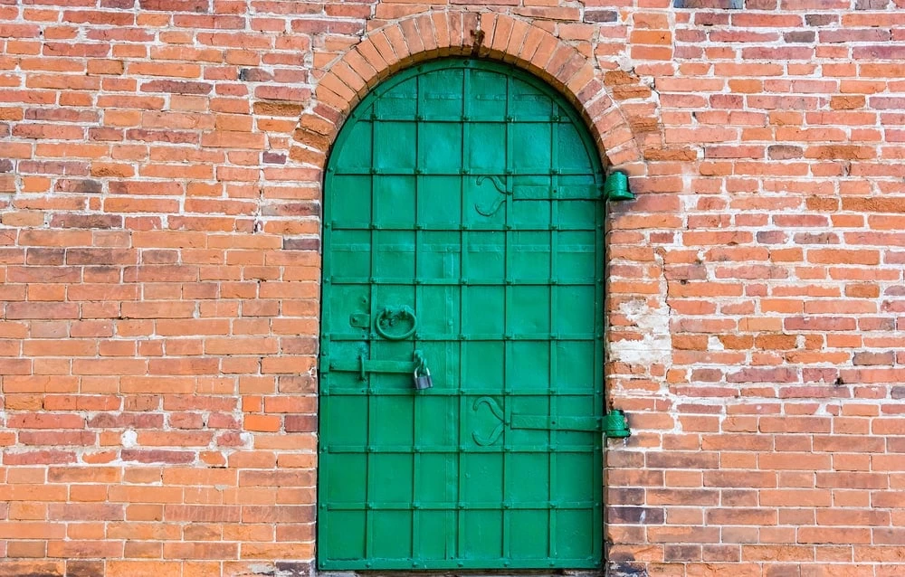 A vibrant green metal door with an arched top is set into a rustic brick wall. The door has a grid pattern, a prominent circular knocker, a long latch, and is secured with a small padlock. A simple bell is mounted on the wall to the right of the door.