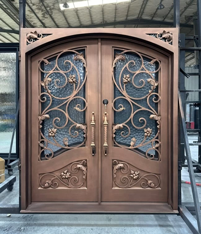 Double doors with an arched top, featuring ornate metalwork of swirling vines and grapes, frosted glass panels, and bronze handles. The doors are set in an industrial workshop environment.