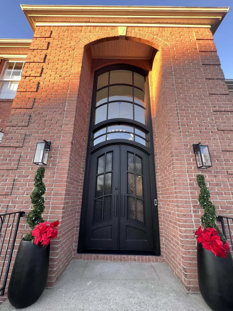 Tall black double doors with arched glass panels set in a brick house entryway. Two black planters with red poinsettias and spiral topiaries flank the entrance; wall-mounted lights are on either side.
