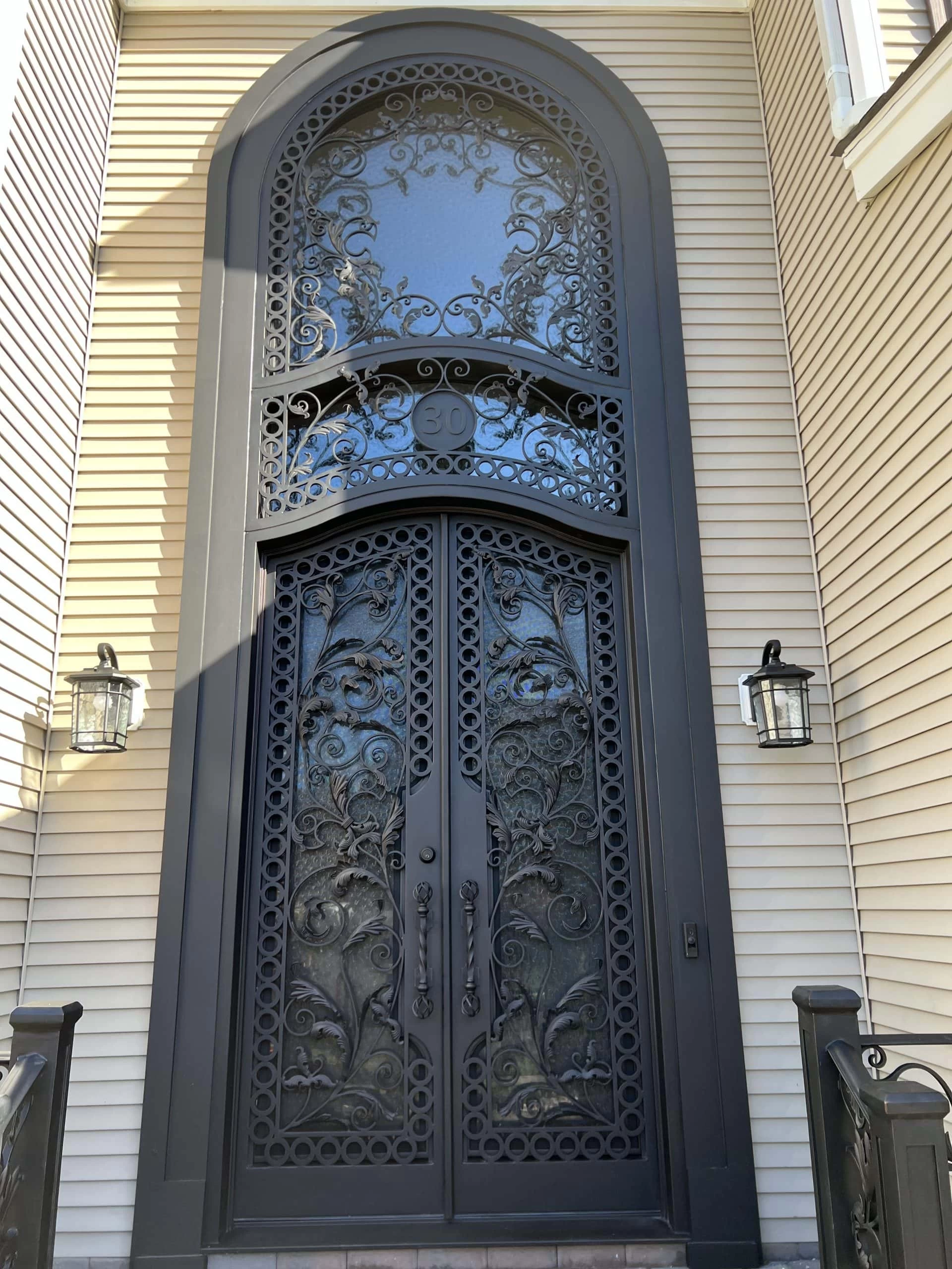 Black ornate double doors with intricate ironwork and decorative glass, topped by an arched transom window. The doors are set in a light-colored exterior wall, flanked by two wall lanterns.