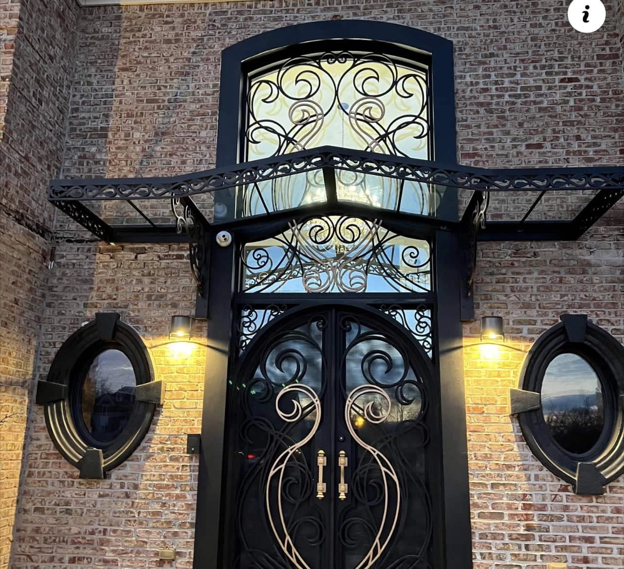 Ornate double black doors with gold swirl designs and matching wrought iron above, set in a brick wall. Two round windows flank the doors, and warm lights illuminate the entrance. A glass and metal awning is above the door.