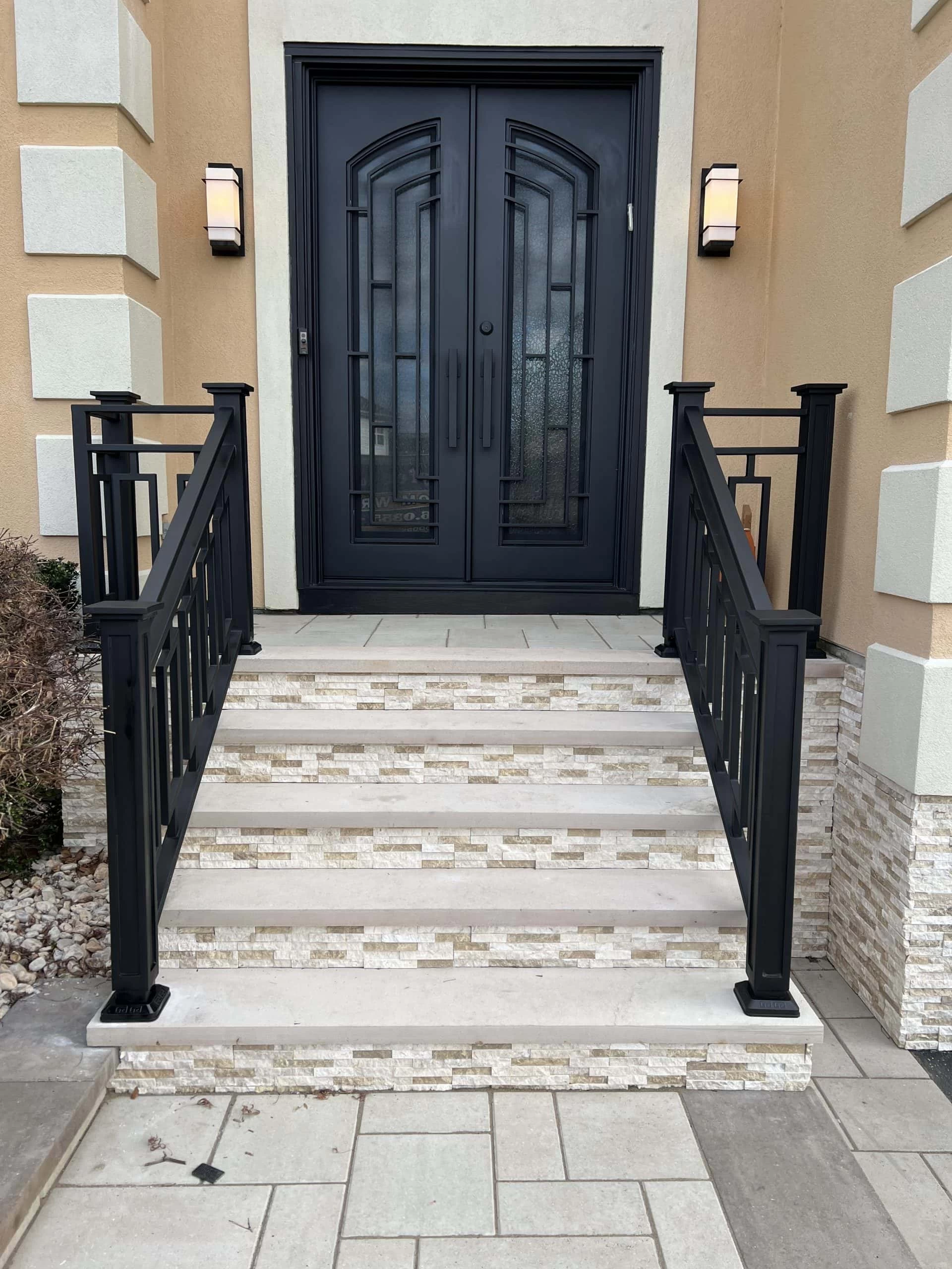 Black double doors with decorative glass panels at the top of a short staircase with beige stone steps and matching black railings. Wall lights are mounted on both sides of the entrance.