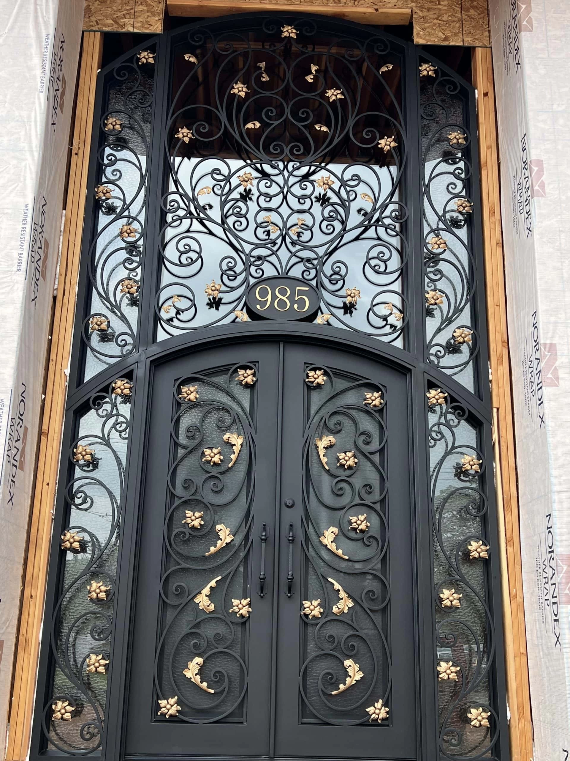 A tall, ornate black metal door with intricate swirling designs and gold floral accents, set in a wooden frame under construction. The address number "985" is displayed above the door.
