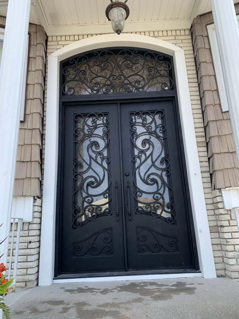A tall black double door with ornate wrought iron scrollwork and glass panels, set in a brick house with tan siding and white trim. A lantern hangs above the arched doorway.