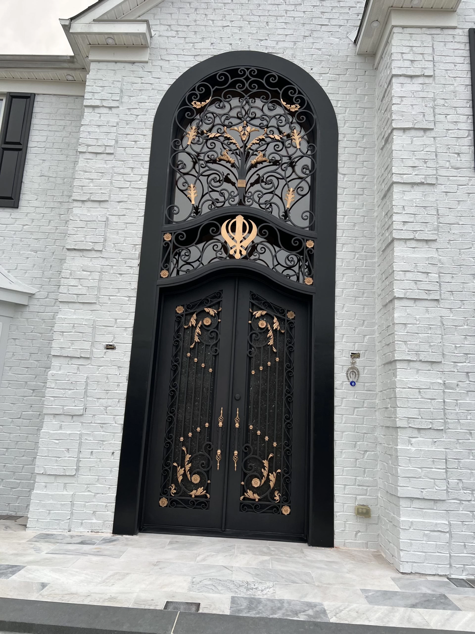 A tall, ornate black door with gold accents stands against a white brick building. Above the door is an arched ironwork design featuring a decorative tree with gold leaves and scroll patterns.