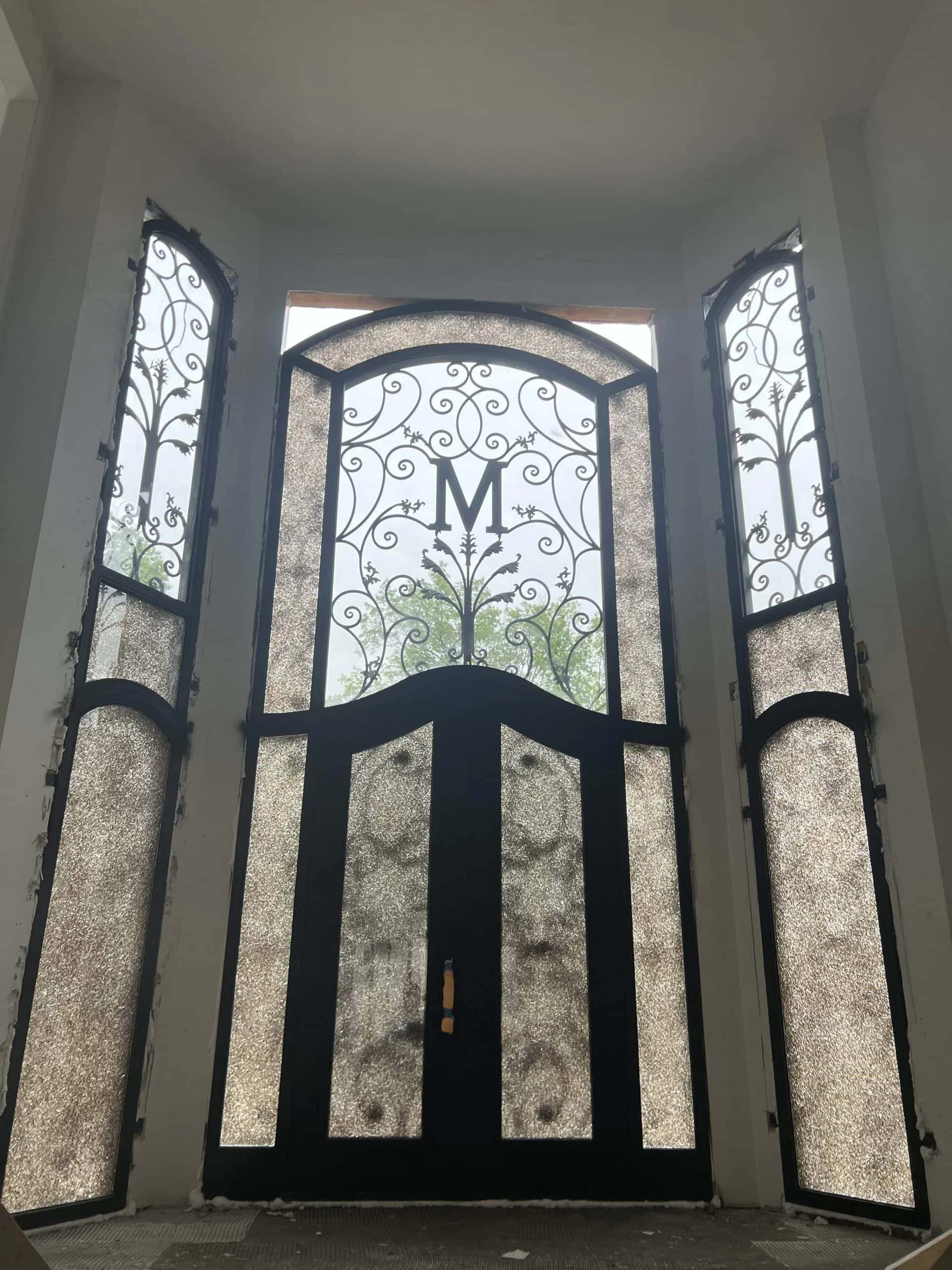 Tall, ornate black double doors with frosted glass panels and intricate ironwork, featuring a large letter "M" in the center, flanked by matching sidelights and transom windows.