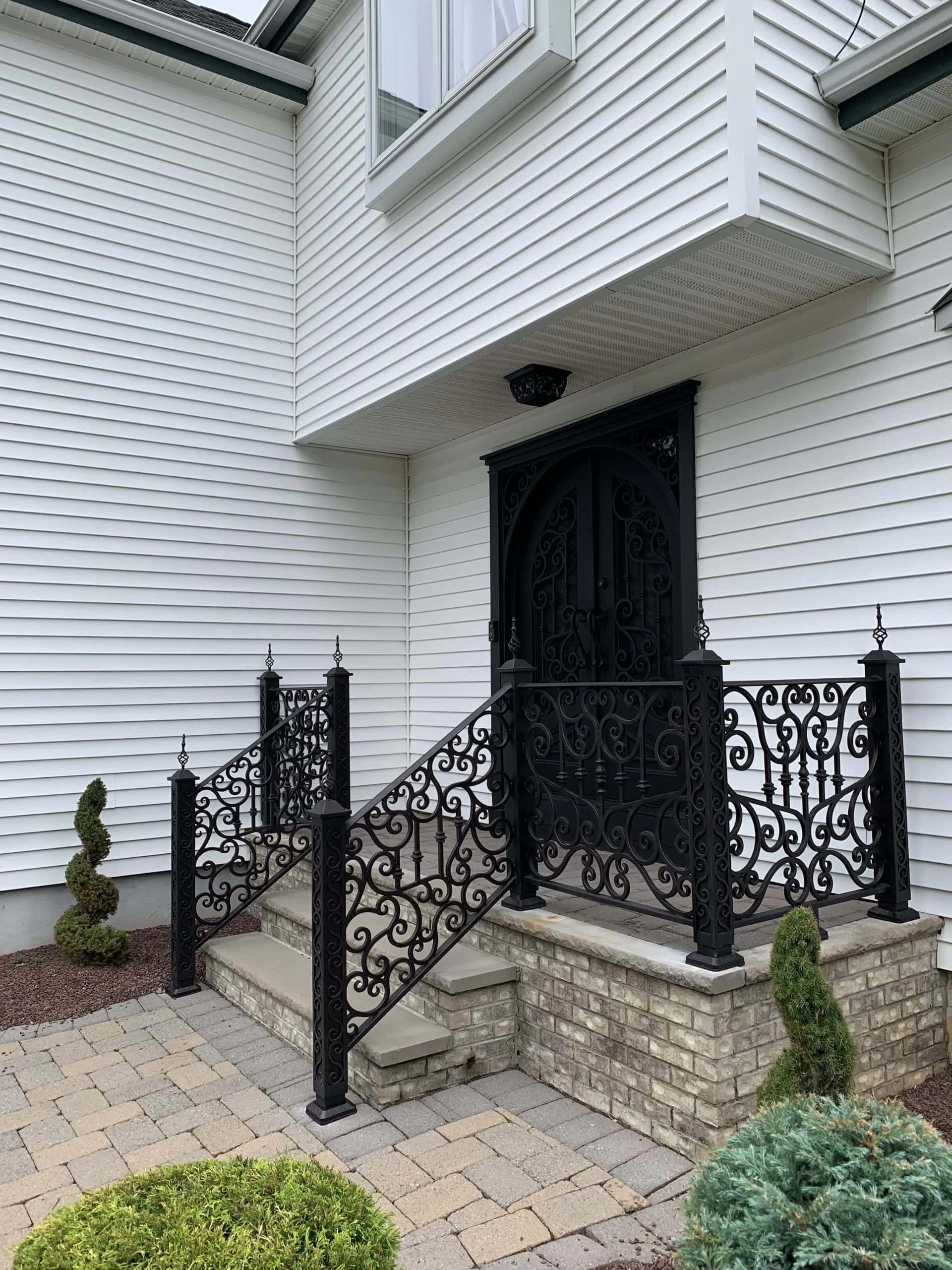 A small porch with decorative black wrought iron railings and gate leads to a double arched black front door on a white house with brick steps and landscaped bushes.