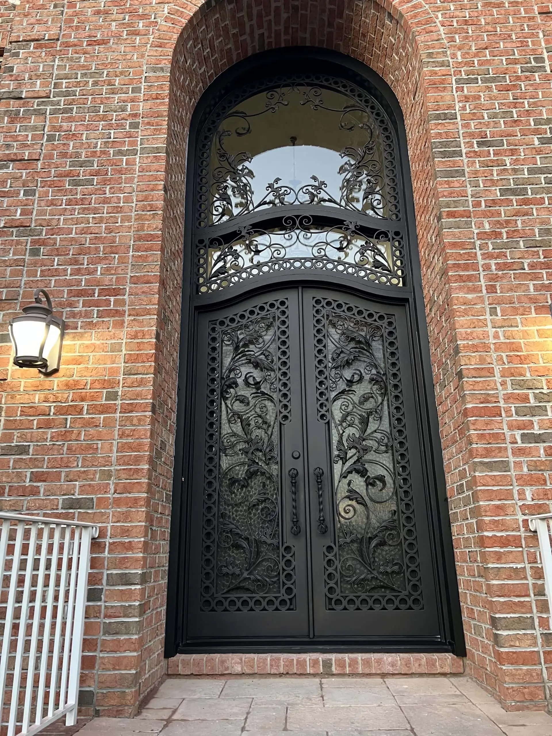 Ornate black double doors with intricate wrought iron designs and glass panels, set in a tall arched brick entryway; a lantern-style light fixture is mounted on the left wall.