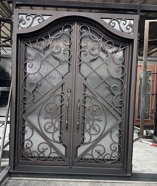 Ornate double iron doors with intricate scrollwork and geometric patterns, featuring glass panels behind the metal design, set in an industrial workshop environment.