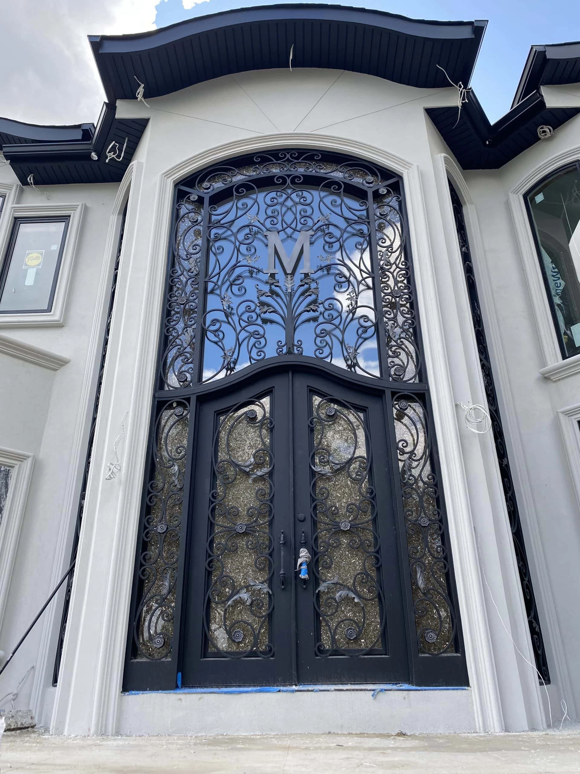 Elegant black double doors with intricate, ornate ironwork and glass panels, topped by a large window featuring more decorative ironwork and the letter "M" at the center. The exterior is light-colored with modern trim.