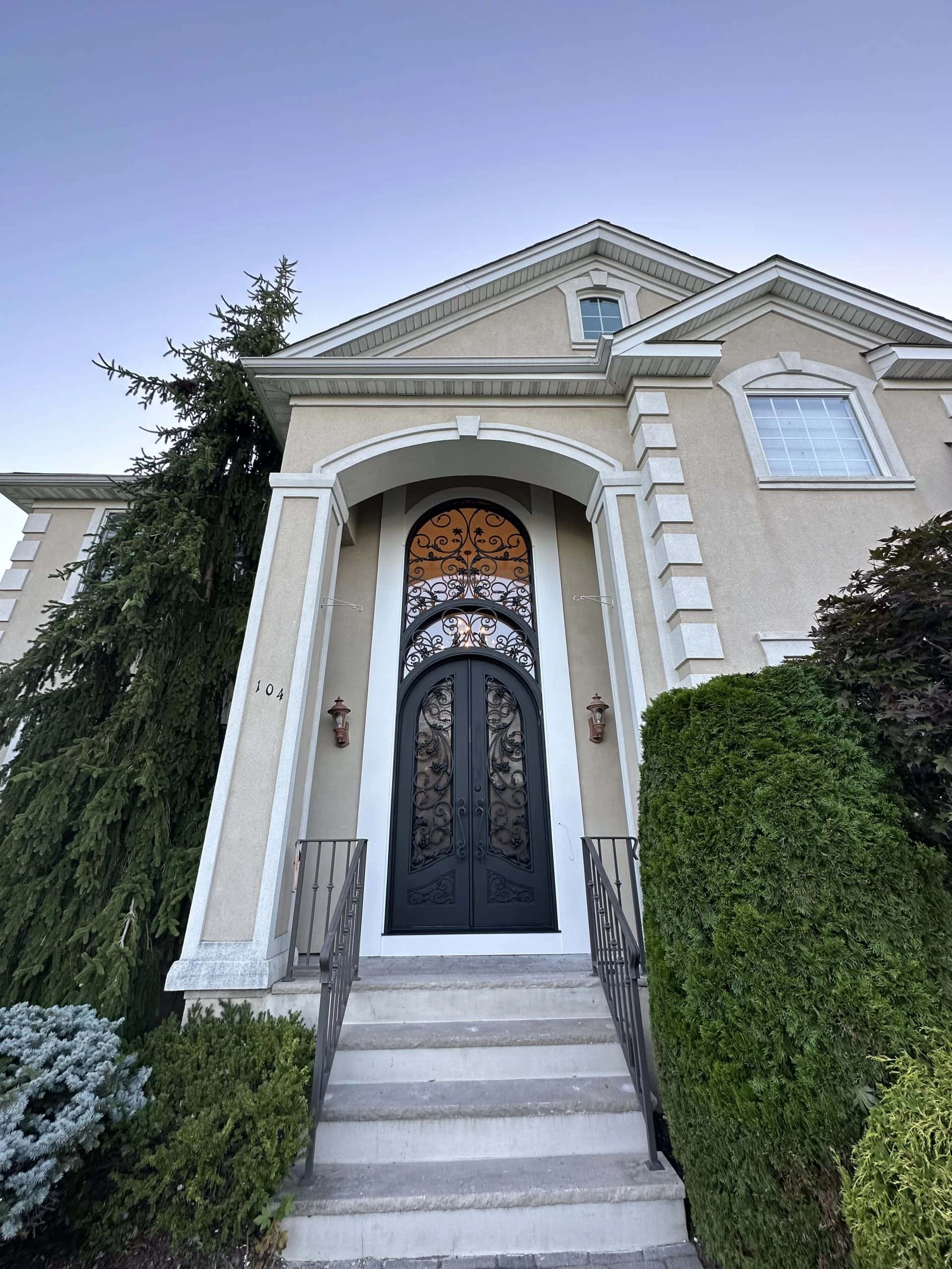 A two-story beige house with a tall arched doorway featuring an ornate black wrought iron door, white trim, and decorative columns. Bushes and a large tree frame the entrance. Steps with railings lead up to the door.