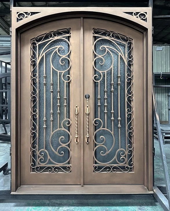 Double bronze-colored metal doors with ornate scrollwork and vertical bars, featuring two large handles and textured glass panels behind the decorative design, set in an arched frame inside a workshop.