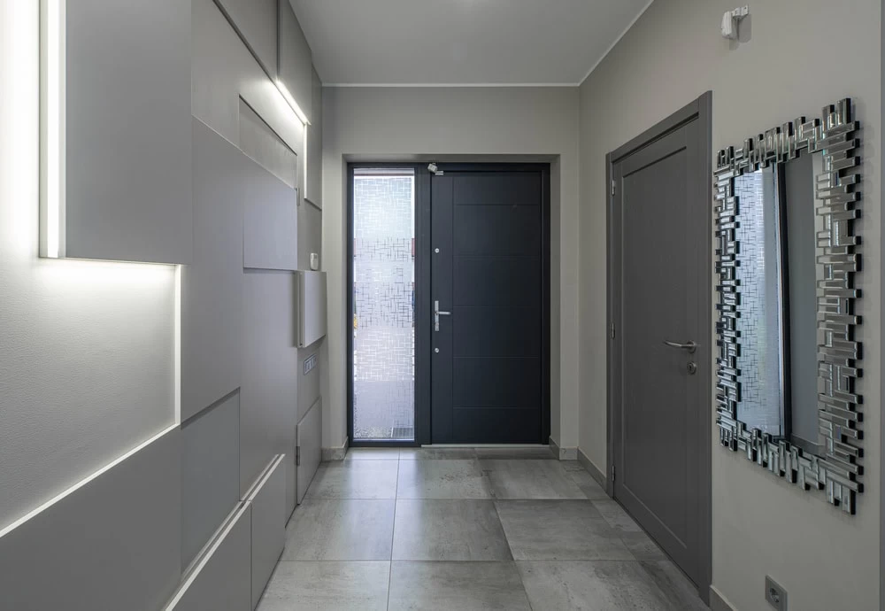 Modern entryway with gray walls and large floor tiles, featuring a custom iron door with sidelights—an ideal choice for homes in Middlesex & Bergen County, NJ—a geometric mirror on the right, and minimalist light panels on the left.