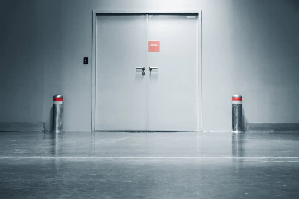 Double gray iron doors with a red sign in the center, flanked by two short posts with red bands, stand in a clean, empty hallway with polished concrete floors and cool lighting—common in NJ’s Middlesex & Bergen County buildings.