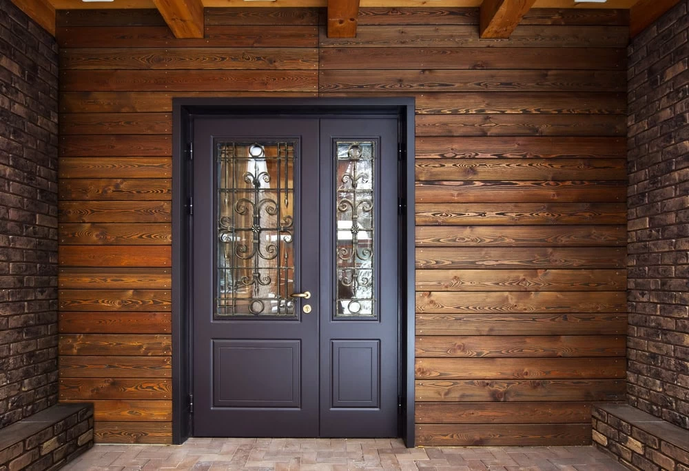 A modern black double door with decorative glass panels and custom ironwork, perfect for homes in NJ. Set in a wooden plank wall with brick accents on either side and a tiled floor, ideal for Middlesex & Bergen County residences.