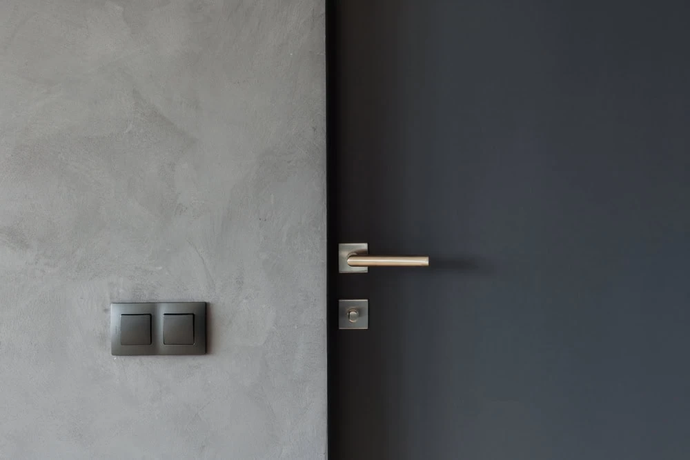 A modern, minimalistic interior featuring a gray textured wall with two black light switches and a sleek, dark-colored custom iron door with a metallic handle—ideal for homes in Middlesex & Bergen County, NJ.