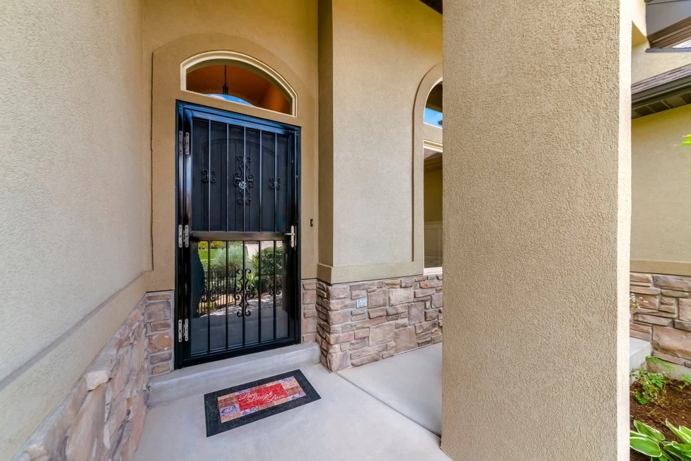 A black metal security door with decorative scrollwork, crafted by custom iron doors Middlesex & Bergen County, NJ, is set in a beige stucco exterior with stone trim. A colorful welcome mat lies on the concrete porch in front of the entrance.