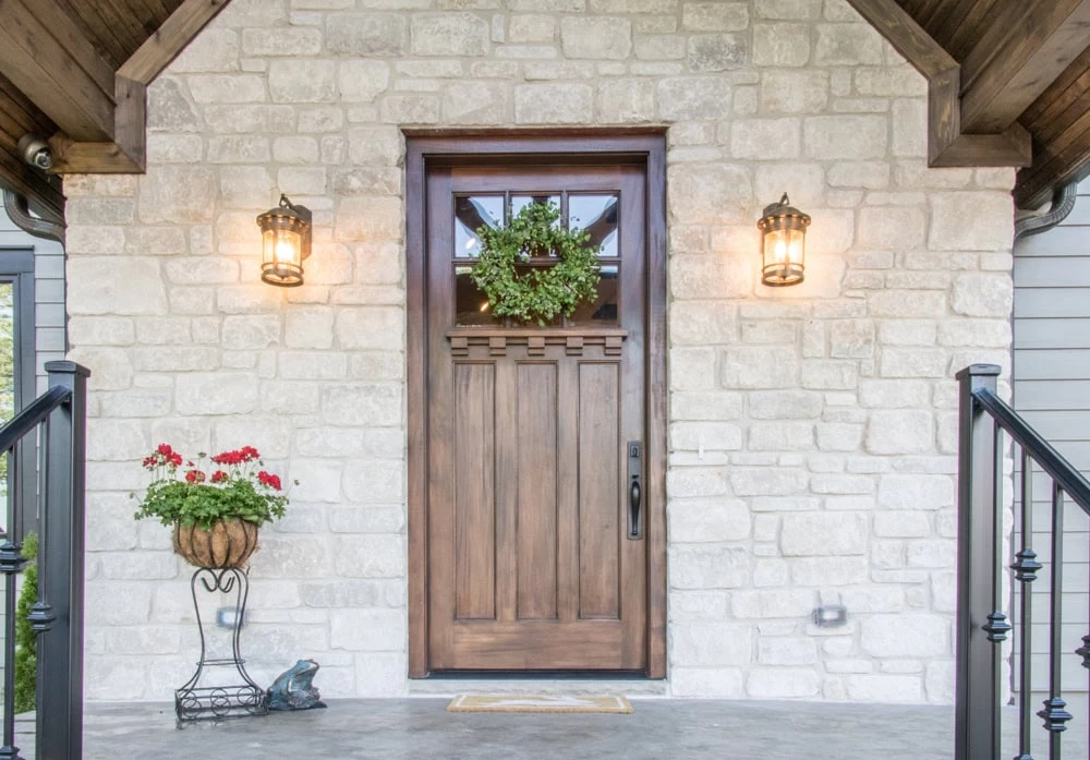 A wooden front door with a green wreath and glass window is set in a stone wall. Two lantern-style lights are on either side, while a planter with red and white flowers adds charm—explore custom iron doors Middlesex & Bergen County, NJ for more options.