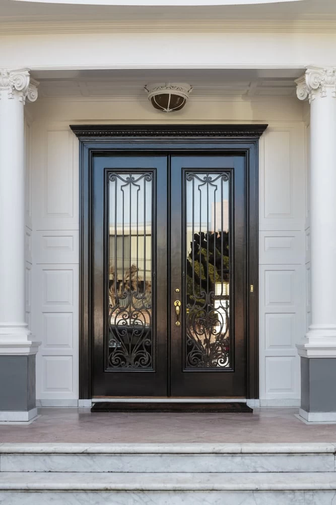 A large, elegant double door with intricate black wrought iron designs by custom iron doors Middlesex & Bergen County, NJ, flanked by white columns and decorative moldings, sits at the top of marble steps in a grand entranceway.