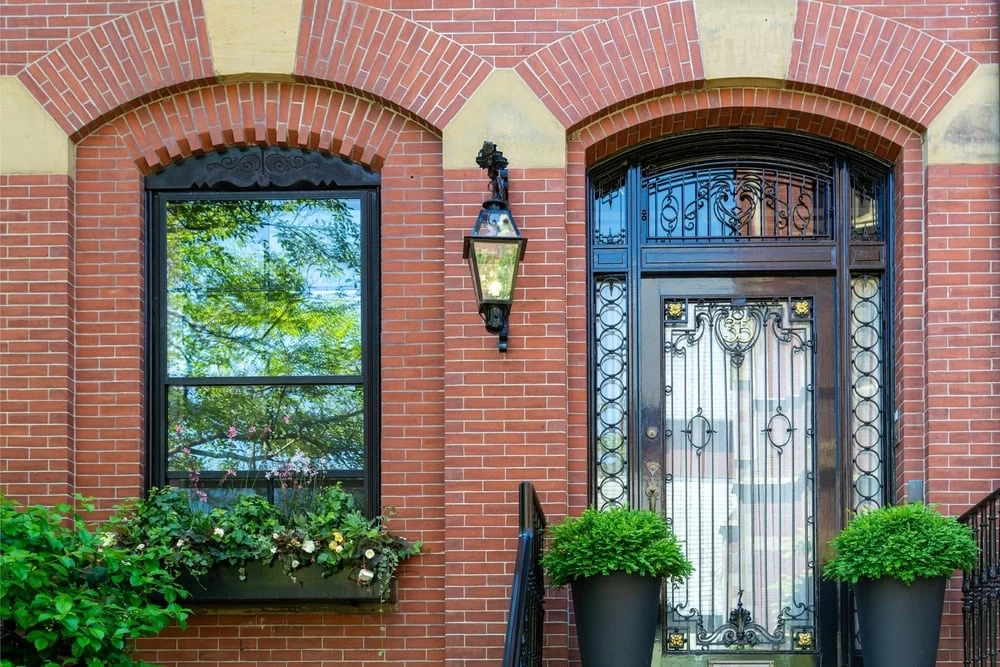 A brick building facade with a black-framed window, a custom iron door with ornate metalwork, lantern-style light, two potted plants by the entrance, and a window box filled with greenery—perfect for homes in Middlesex & Bergen County, NJ.