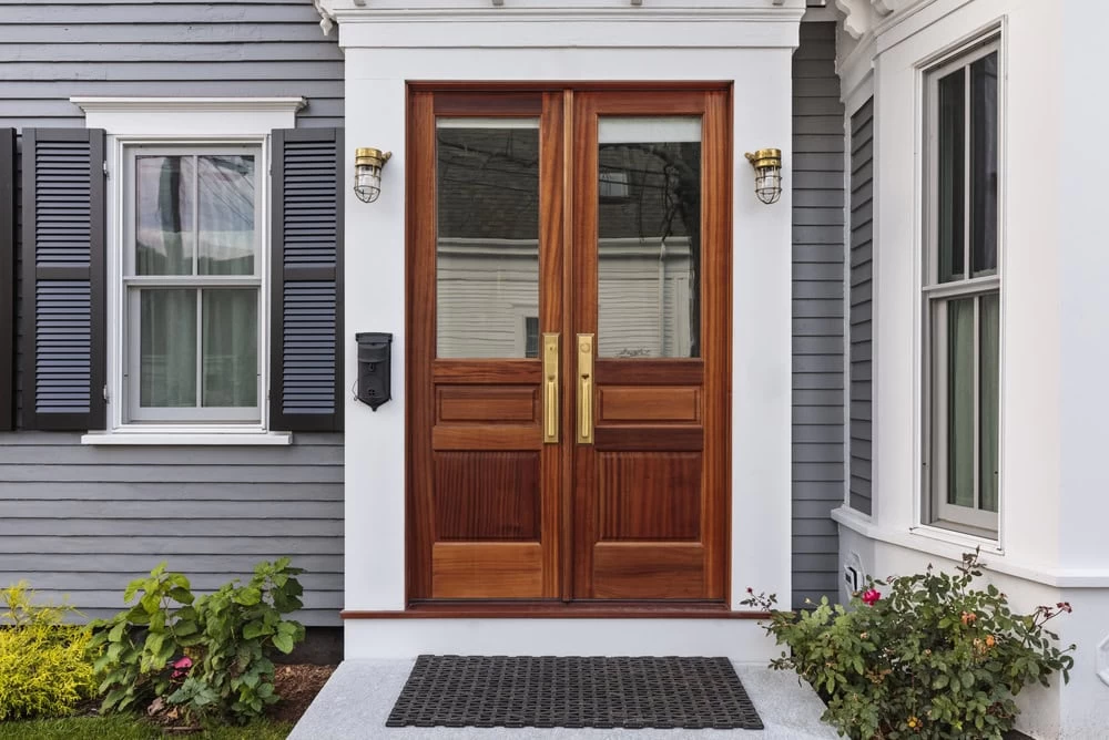 Double wooden front doors with glass panels and brass handles are framed by white trim, set in a gray NJ house with black shutters. Two wall-mounted lights and a black mailbox are on either side, with flowers and a doormat in front.