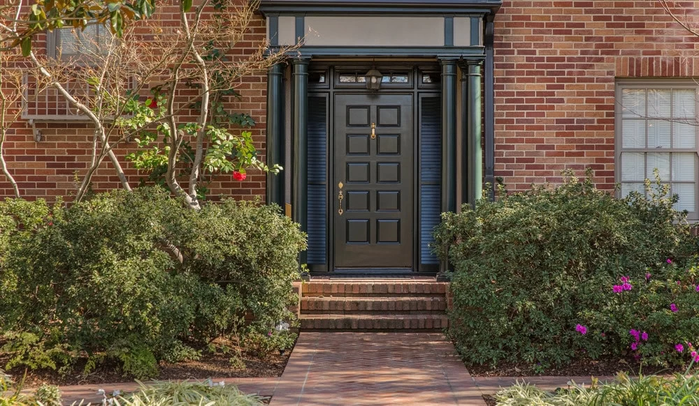 A black custom iron door with brass hardware is centered on a red brick house in NJ, framed by columns and greenery, with a brick path and steps leading up to the entrance—showcasing the elegance of iron doors in Middlesex & Bergen County.