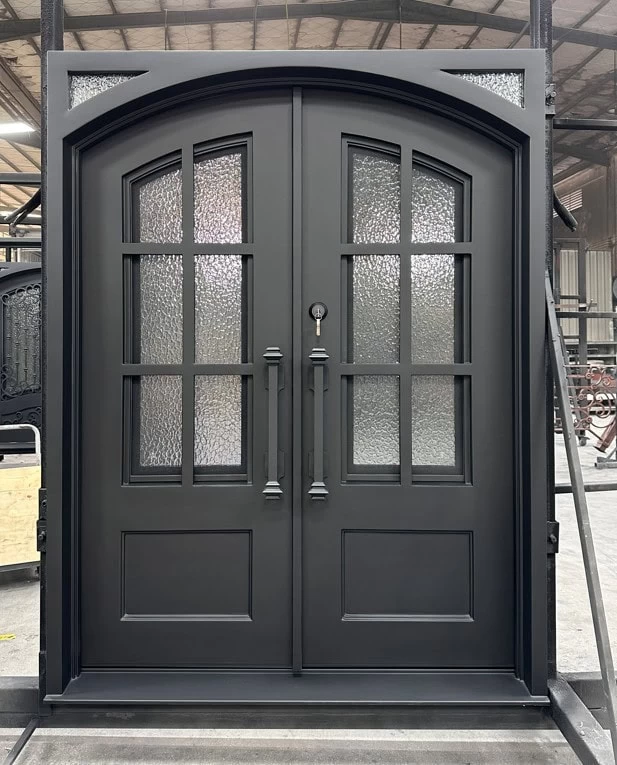 A double black metal door with arched top, decorative glass window panels, and long handles stands in an industrial workshop setting.
