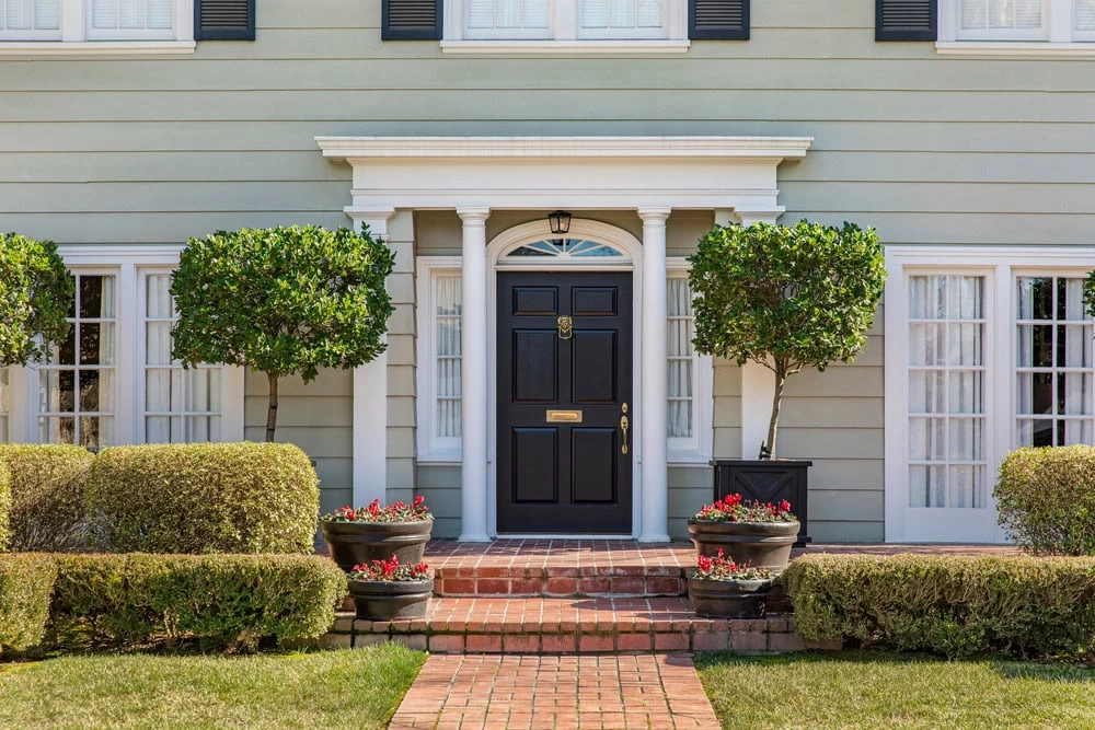 A symmetrical front entry of a house features custom iron doors Middlesex & Bergen County, white columns, two trimmed trees in planters, potted red flowers, brick steps, and manicured hedges.