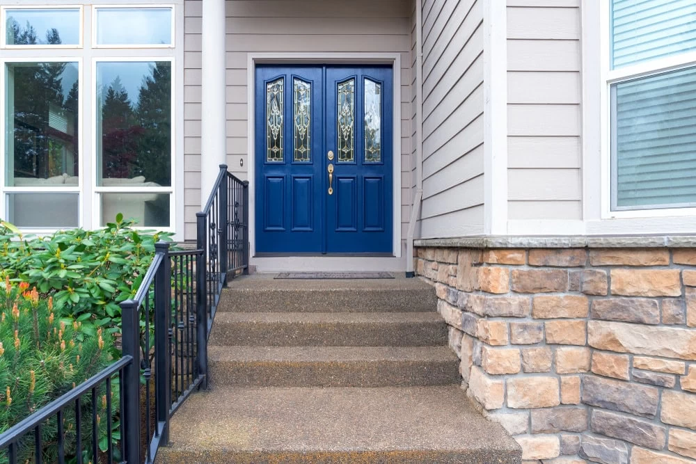 Front entrance of a house with blue double doors, decorative glass panels, stone siding, beige paneling, a small stairway with black railings, and green shrubs—perfect for showcasing custom iron doors Middlesex & Bergen County, NJ.