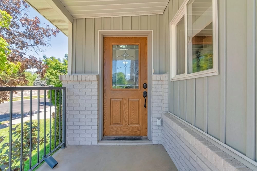 A wooden front door with decorative glass panel is centered on a small porch with light gray brick and siding. Black custom iron doors in Middlesex & Bergen County, NJ, line the edge, with a window on the right side and green trees visible outside.