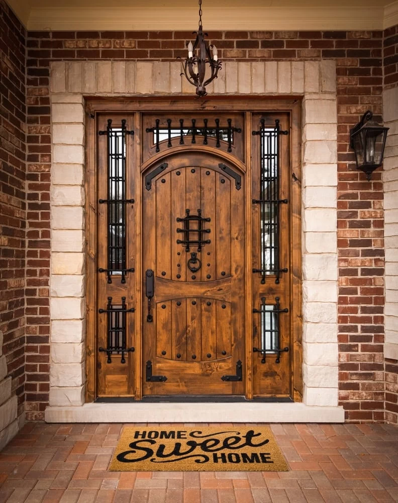 A wooden front door with custom iron doors Middlesex & Bergen County-style decorative ironwork and stone trim, flanked by brick walls. A doormat says “Home Sweet Home,” while a lantern hangs above the NJ entrance.